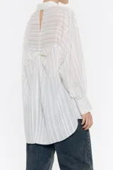 Camisa larga de viscosa con textura, de corte recto, con cuello camisero, manga larga de puño con botón y cierre frontal con abotonadura. Presenta bolsillo de plastrón en el pecho y nudo decorativo en la parte trasera para dar volumen.