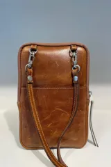 Bolso pequeño tipo bandolera o cartera de mano, confeccionado en cuero vacuno color taupe/marrón claro, con detalles en material metalizado plateado. Presenta un bordado de flor de lis en el frente y cierre superior. Incluye correa fina desmontable.