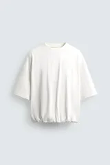 Camiseta blanca de algodón, corte holgado, cuello redondo, manga corta y bajo con elástico interior.