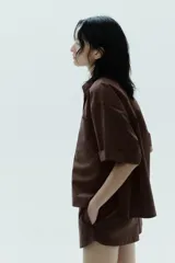 Camisa marrón de popelín con cuello solapa, manga corta con vuelta, bolsillo de plastrón en el pecho, bajo con aberturas laterales, pinzas en la espalda y cierre frontal con botones ocultos.