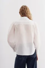 Camisa blanca de algodón con corte recto, cuello clásico y cierre frontal con botones. Presenta detalles de puntilla en cuello y cartera, mangas largas abullonadas con puños elastizados.
