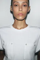 Camiseta blanca de algodón con cuello redondo, manga corta con dobladillo y dos bolsillos de parche en el frente con detalle de aplicación metálica.