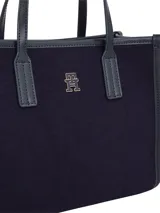 Cartera tipo tote bag de estructura rígida, color azul marino, con cuerpo principal de material textil tipo fieltro y ribetes y asas de poliuretano liso. Presenta un pequeño logo metálico TH en la parte frontal superior y un colgante decorativo con el escudo de la marca.