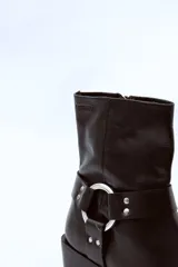 Bota de cuero negra con caña media, puntera ligeramente afilada y detalle de correa con herraje metálico circular y tachas. Taco bajo y ancho.