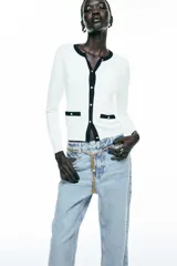Chaqueta blanca de punto con cuello redondo y manga larga. Tiene falsos bolsillos de vivo en el delantero y cierre frontal con botones dorados.