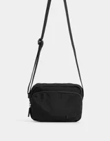 Bandolera negra de tela deportiva con bolsillo frontal con cierre, correa regulable y tag de goma.