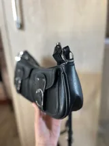 Cartera bandolera negra de cuero sintético, con correa ajustable y dos bolsillos frontales con hebillas plateadas.