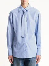 Camisa de manga larga, corte holgado, con cuello camisero y estampado de rayas diplomáticas verticales en blanco y azul. Incluye una corbata ancha integrada en el mismo tejido y patrón, anudada al cuello.