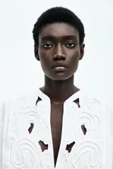 Blusa blanca de algodón con escote en V y pechera con broderie tono sobre tono embellecido con minicuentas y ribetes con pompones. Modelo de corte holgado con hombros caídos y mangas largas con tapeta de manga y puños abotonados. Bajo redondeado.