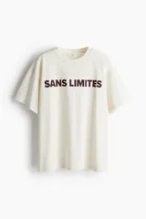 Remera color crema de corte holgado con cuello redondo y hombros caídos. Estampado frontal con la frase "Sans Limites" en color marrón.