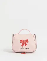 Neceser de viaje color rosa con cierre superior y manija. Estampado frontal con un moño rojo con corazones y la frase "Très Chic".