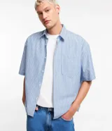 Camisa masculina oversized boxy de manga corta, confeccionada en tejido oxford a rayas verticales celestes y blancas. Cuenta con cuello inglés, abotonadura frontal y bolsillo de parche en el pecho.