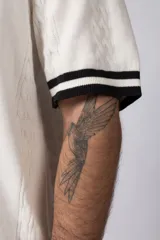 Camisa tejida color crudo, de manga corta, con cuello bowling y botones frontales. Presenta un tejido con textura de trenzas verticales y rib negro en los puños.