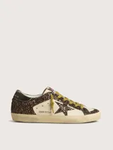 Zapatillas deportivas Golden Goose modelo Super-Star, con cuerpo principal de cuero color crema y detalles de glitter marrón oscuro. Presentan la estrella lateral y el talón en glitter marrón oscuro, cordones amarillos y suela desgastada.