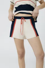 Short tejido en jersey color crudo con cintura elastizada azul marino y cordón rojo. Presenta rayas verticales rojas y laterales en azul marino.