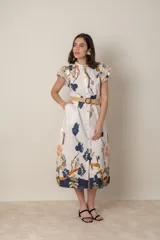 Vestido midi de algodón color natural con estampado floral en tonos azules, naranjas y marrones. Tiene cuello mao, mangas cortas con volados, bolsillos laterales y cinturón de soga color beige.