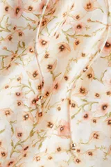 Blusa de tejido crepé color crema con estampado de flores pequeñas en tonos rosados y marrones. Tiene corte holgado, cuello con volados y escote en V con tiras para ajustar. Las mangas son cortas tipo mariposa con ribete de volado.