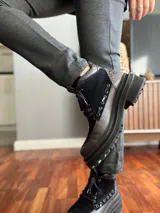 Borcego de cuero negro con puntera redondeada, cordones negros, paneles laterales de gamuza negra y detalle de cuero texturizado en el tobillo. Suela track de goma negra.