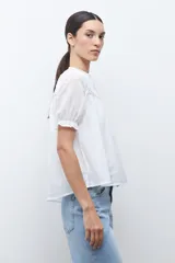 Camisa blanca de manga corta con cuello mao y detalles de bordado en el canesú superior. Presenta frunces en el pecho y la espalda, y un cierre frontal de botones.