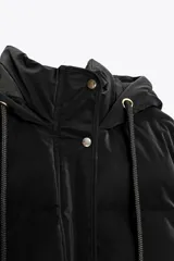 Anorak acolchado color verde oliva, resistente al viento y con aislamiento térmico. Tiene cuello alto con capucha ajustable con cordones, manga larga con puño interior, bolsillos delanteros con forro polar y cierre de cremallera, y cierre frontal con cremallera oculta y botones a presión.