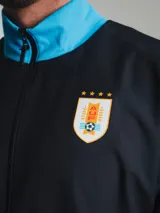 Campera deportiva negra con detalles celestes, cierre frontal y cuello alto. Logo de Nike bordado en el pecho y escudo de Uruguay en el brazo.