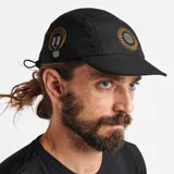 Gorro tipo camper negro con textura microperforada, visera corta y construcción plegable. Presenta un bordado frontal de un sol estilizado en tonos naranjas y un bordado lateral de una serpiente con una hoja en tonos amarillos y blancos.