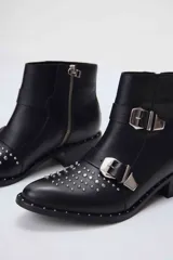 Botas cortas de cuero negro con tachas plateadas en la puntera y el talón, cierre lateral con cremallera y hebillas decorativas. Taco cuadrado bajo.