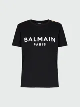 Remera negra de algodón con cuello redondo y estampado del logo de Balmain en blanco. Tiene botones dorados decorativos en el hombro.