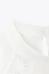 Blusa blanca de cuello redondo y manga larga con puños. Presenta bordados florales y entredoses combinados en el frente. Cierre en la espalda con abertura y botón.