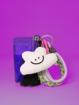 Llavero con tarjeta identificadora transparente con borde violeta, dije de nube blanca con cara sonriente, borla de hilos negros, mosquetón y argolla violeta, cinta de soga trenzada con diseño geométrico en amarillo, violeta y blanco.