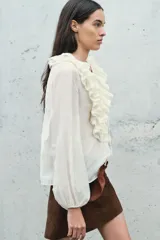 Blusa color crudo confeccionada en hilatura con mezcla de lyocell. Escote pico ajustable con cordones, manga larga con volumen acabada en elástico y detalle de volantes en el mismo tejido.