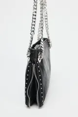 Bolso alargado negro con tachas plateadas en el borde inferior y detalle de cremallera en el corte. Tiene asa de cadena plateada multiposición para usar al hombro o como bandolera. Incluye bolsillo interior con cremallera y cierre principal con cremallera.