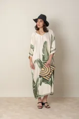 Vestido largo color natural con estampado de hojas de palmera en tonos verdes. Tiene corte amplio tipo túnica, cuello redondo, mangas tres cuartos, bolsillos laterales y ruedo con ribete.