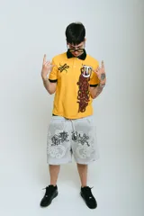 Bermuda de felpa color gris claro con efecto lavado (washed). Presenta un corte ancho con cintura elástica y cordón beige. El diseño frontal incluye un estampado serigrafiado en negro con texto y motivos florales/ornamentales en bordado blanco. Posee remaches metálicos cromados en los laterales y bolsillos.