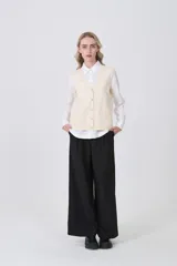 Chaleco de punto color crema, con cuello en V, cierre frontal de botones y dos bolsillos frontales.