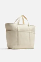 Bolso tipo shopper color crema, fabricado en tejido de algodón. Tiene doble asa de mano, compartimento principal con cierre de cremallera, seis bolsillos planos interiores, funda interior para dispositivos electrónicos de hasta 13 pulgadas con cierre de cremallera, bolsillo plano frontal y bolsillos planos laterales.
