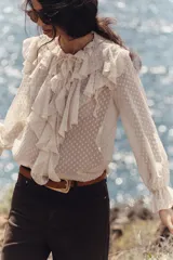 Blusa color crema semitransparente con cuello redondo con volantes y cordón. Manga larga acabada en elástico y volante. Cierre frontal con botones.