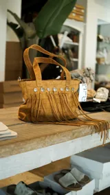 Bolso de cuero color beige con doble asa corta y correa larga extraíble. Presenta detalle de flecos largos en la parte frontal y herrajes metálicos plateados. Interior forrado con bolsillos. Marca Brava.