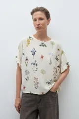 Blusa de manga corta, color crudo, con estampado floral en tonos azules, amarillos y rojos. Presenta cuello redondo y volados en los puños.