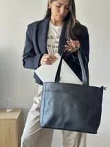 Cartera tipo tote de cuero graneado color negro, con textura uniforme y suave al tacto. Tiene manijas de 55 cm, bolsillo frontal con broche imantado, bolsillo trasero con cierre metálico, cierre principal metálico, forro interior y tres bolsillos internos (uno con cierre, uno plaqué y uno tipo sobre con solapa y botón).