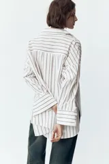 Camisa blanca con rayas verticales grises, de corte holgado, cuello solapa y mangas largas con puños abotonados. Tiene un bolsillo de parche en el pecho, pinzas en la espalda y bajo asimétrico con aberturas laterales.