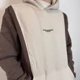 Canguro con capucha color marrón y beige, con logo bordado en el pecho y bolsillo tipo canguro.