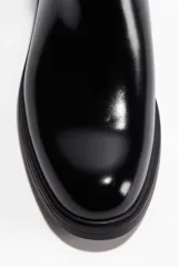 Botas Chelsea negras de cuero sintético con acabado brillante, elásticos laterales y trabillas de tela en el talón y la parte delantera. Suela de goma gruesa y plana.