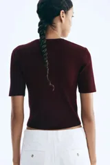 Cárdigan corto de punto acanalado en color vino tinto, con cuello redondo, manga corta y cierre frontal de botones dorados. Presenta dos bolsillos delanteros decorativos con botón dorado a la altura de la cadera.