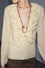 Camisa color beige de algodón con escote en V y volantes. Tiene mangas largas con botón en los puños y cierre con lazos en el frente.