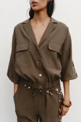 Camisa marrón de cuello solapa y manga corta con trabilla y botón. Tiene bolsillos delanteros de plastrón con solapa, bajo elástico y cierre frontal con botones a presión.