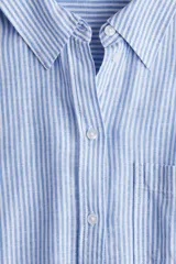 Camisa celeste y blanca a rayas verticales, confeccionada en mezcla de lino y viscosa. Tiene cuello clásico, botones frontales, bolsillo de parche en el pecho, mangas largas con puños abotonados y bajo redondeado.
