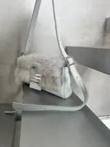 Cartera mini de cuero blanco con solapa de pelo gris y herrajes metálicos plateados.