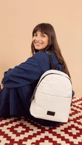 Mochila de tela color blanco con diseño capitoneado en la parte superior y textura tipo telar en la parte inferior. Tiene correas negras ajustables, asa superior y bolsillo frontal amplio. Incluye compartimento interno para iPad.