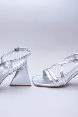 Sandalias de tacón alto color violeta metalizado, con tiras finas cruzadas en el empeine, puntera cuadrada y cierre con hebilla en el tobillo. El tacón es geométrico con forma de triángulo invertido.